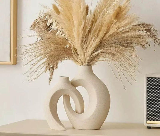 Vases cream ceramic sculptural pair on a shelf in a living room, with dried pampas grass beside a small speaker.